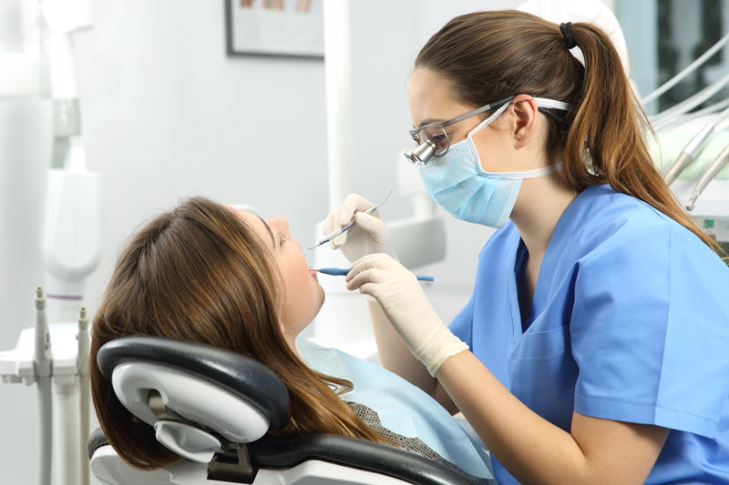 Dentist wearing eyeglasses gloves and mask examining a patient tooth with a dental probe and a mirror.